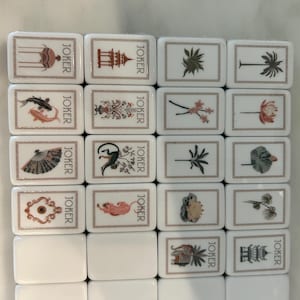 May include: A set of white rectangular mahjong tiles arranged in a grid. Each tile features a thin brown border and the word JOKER printed vertically. The tiles display various illustrations including a pagoda, fish, fan, monkey, tiger, and elephant.