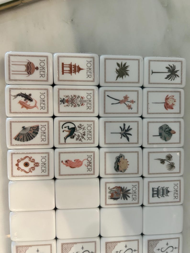 May include: A set of white rectangular mahjong tiles arranged in a grid. Each tile features a thin brown border and the word JOKER printed vertically. The tiles display various illustrations including a pagoda, fish, fan, monkey, tiger, and elephant.