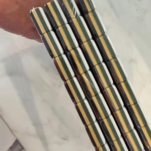 May include: A stack of four rectangular tiles wrapped in clear plastic. Each tile features a repeating horizontal striped pattern consisting of alternating bands of dark brown, white, olive green, and a mustard yellow color throughout the surface.