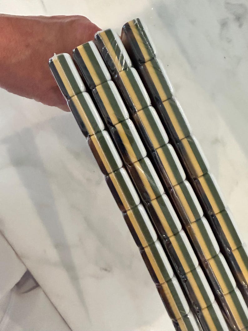 May include: A stack of four rectangular tiles wrapped in clear plastic. Each tile features a repeating horizontal striped pattern consisting of alternating bands of dark brown, white, olive green, and a mustard yellow color throughout the surface.