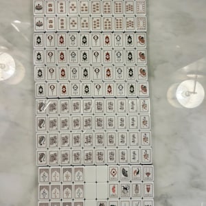 May include: A complete set of rectangular white mahjong tiles arranged in a grid. The tiles feature various black, red, and green engraved designs including numbers, Chinese characters, directional symbols, and illustrated joker tiles with text.