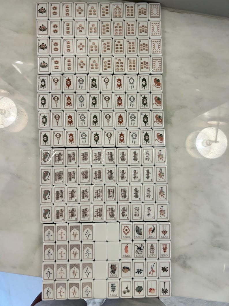 May include: A complete set of rectangular white mahjong tiles arranged in a grid. The tiles feature various black, red, and green engraved designs including numbers, Chinese characters, directional symbols, and illustrated joker tiles with text.