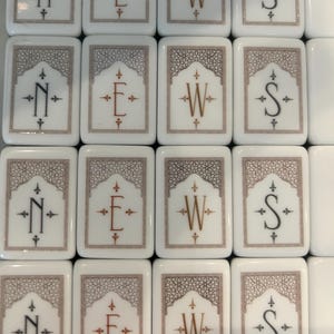 May include: A collection of rectangular white tiles arranged in a grid. Each tile features a central letter N, E, W, or S in a decorative font, surrounded by a geometric border pattern in a muted brown tone with small cross-shaped accents.