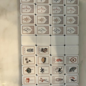 May include: A set of rectangular white Mahjong tiles arranged in a grid. The tiles feature engraved designs including compass directions N, E, W, S, floral motifs, palm trees, and Joker tiles with various illustrations and the text JOKER printed on them.