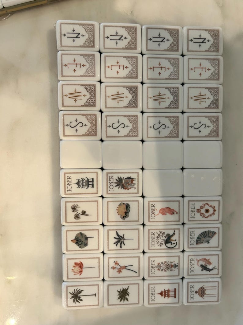 May include: A set of rectangular white Mahjong tiles arranged in a grid. The tiles feature engraved designs including compass directions N, E, W, S, floral motifs, palm trees, and Joker tiles with various illustrations and the text JOKER printed on them.