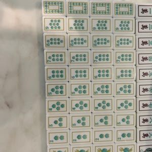 May include: A collection of white rectangular Mahjong tiles arranged in a grid. The tiles feature green floral patterns, numerical indicators, and black Chinese characters. Each tile has a smooth, glossy finish with gold-coloured border accents.