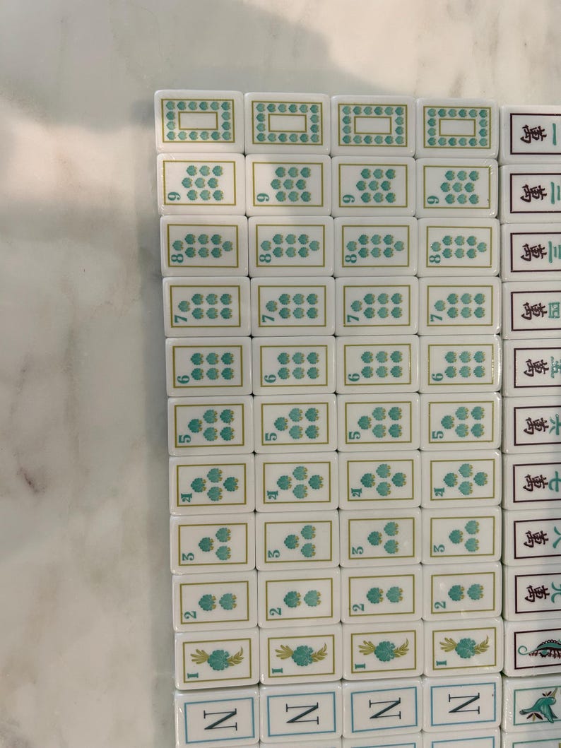 May include: A collection of white rectangular Mahjong tiles arranged in a grid. The tiles feature green floral patterns, numerical indicators, and black Chinese characters. Each tile has a smooth, glossy finish with gold-coloured border accents.
