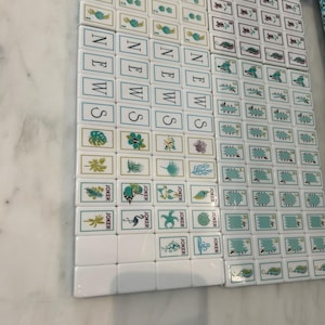 May include: A set of white Mahjong tiles featuring various green and blue illustrations, including floral patterns, sea life, and the word NEWS printed vertically in blue. The tiles are arranged in a grid, with some marked as JOKER tiles.