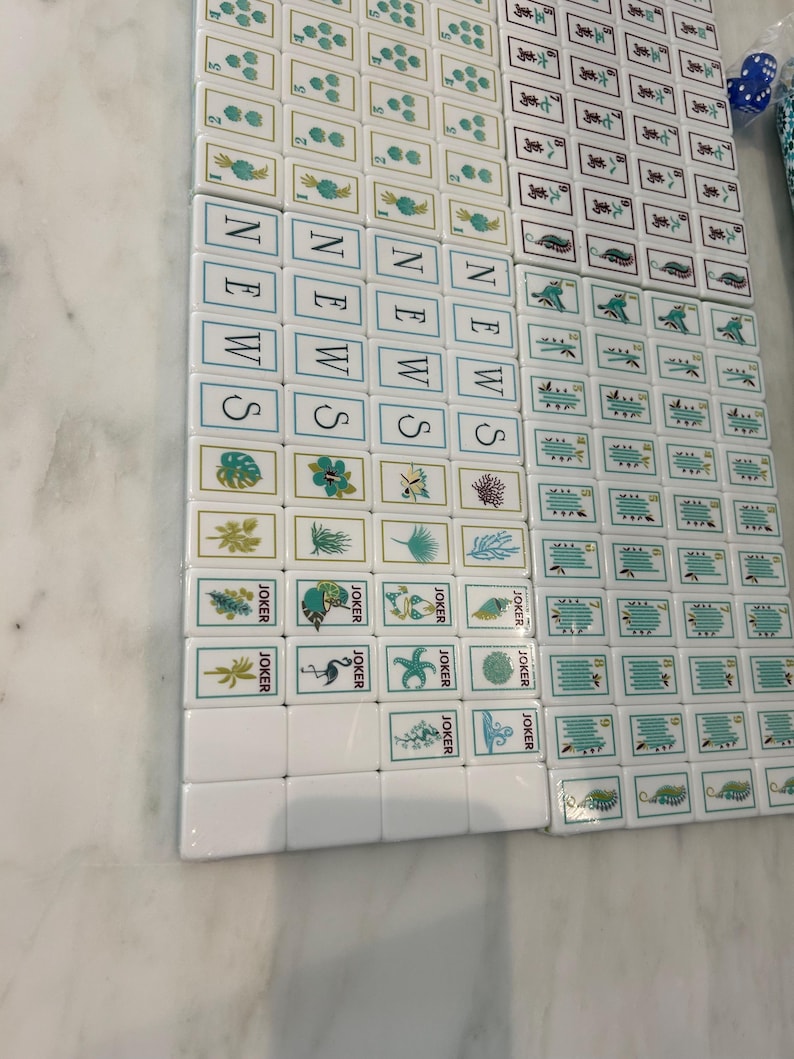 May include: A set of white Mahjong tiles featuring various green and blue illustrations, including floral patterns, sea life, and the word NEWS printed vertically in blue. The tiles are arranged in a grid, with some marked as JOKER tiles.