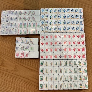 May include: A collection of white rectangular mahjong tiles arranged in groups. The tiles feature colourful illustrations including hot air balloons, matryoshka dolls, floral patterns, bamboo stalks, birds, and various numbered and lettered designs.