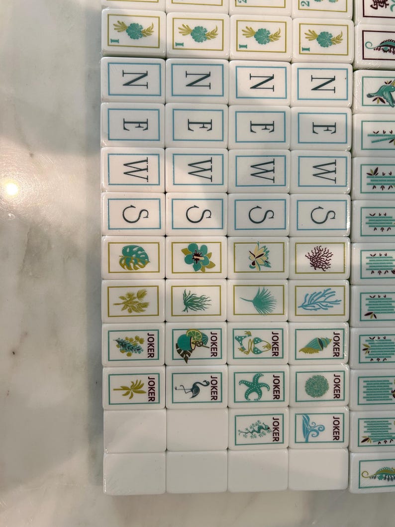 May include: A collection of white rectangular Mahjong tiles arranged in rows and columns. The tiles feature various colourful illustrations including tropical plants, sea life, and the word NEWS printed vertically in black letters, alongside JOKER tiles.