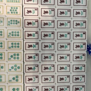 May include: A collection of white rectangular Mahjong tiles arranged in rows. The tiles feature green floral patterns, black Chinese characters, and Arabic numerals. Some tiles display intricate green and black designs at the bottom edges.