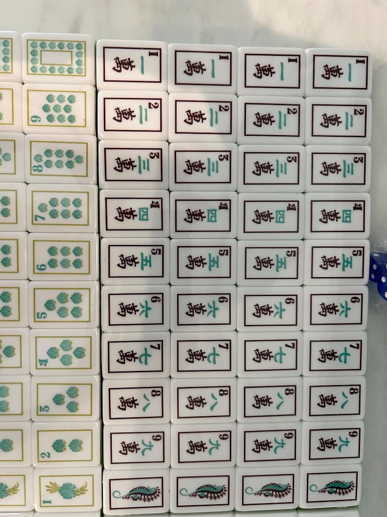 May include: A collection of white rectangular Mahjong tiles arranged in rows. The tiles feature green floral patterns, black Chinese characters, and Arabic numerals. Some tiles display intricate green and black designs at the bottom edges.