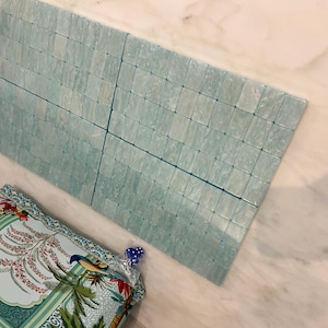 May include: A rectangular grid of light blue, translucent, square-shaped tiles with a marbled texture. Below the tiles, a portion of a patterned fabric is visible, featuring a colourful illustration of a bird, palm leaves, and floral motifs.