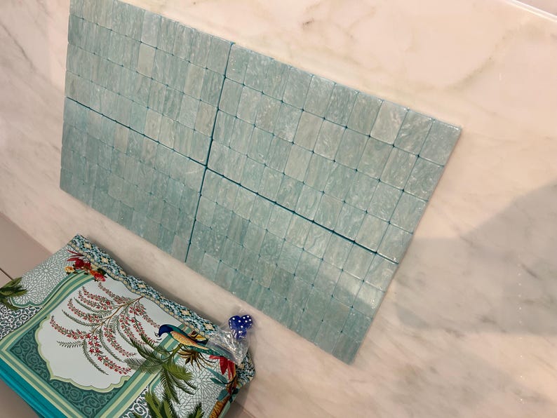 May include: A rectangular grid of light blue, translucent, square-shaped tiles with a marbled texture. Below the tiles, a portion of a patterned fabric is visible, featuring a colourful illustration of a bird, palm leaves, and floral motifs.