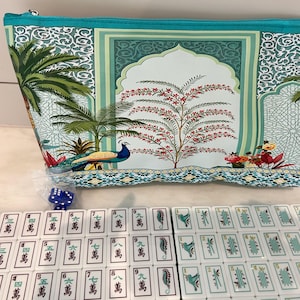 May include: A teal zippered pouch featuring a printed design of palm trees, a peacock, and an arched architectural motif. Below the pouch are rows of white mahjong tiles with green and black characters and symbols, plus two small blue dice.