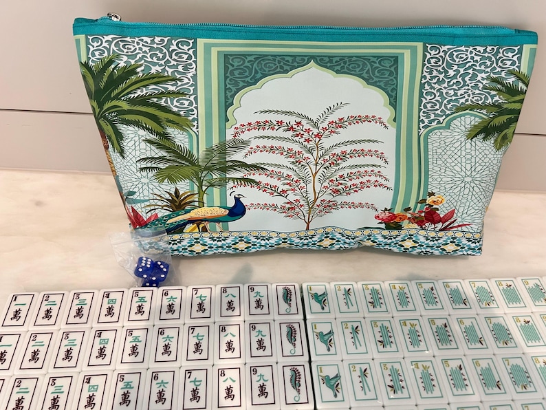 May include: A teal zippered pouch featuring a printed design of palm trees, a peacock, and an arched architectural motif. Below the pouch are rows of white mahjong tiles with green and black characters and symbols, plus two small blue dice.