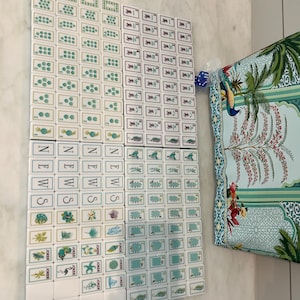 May include: A set of white mahjong tiles featuring green and black ink illustrations of numbers, symbols, and nature motifs, including tiles labelled NEWS and JOKER. A blue die and a patterned fabric bag with a peacock and tree design are included.