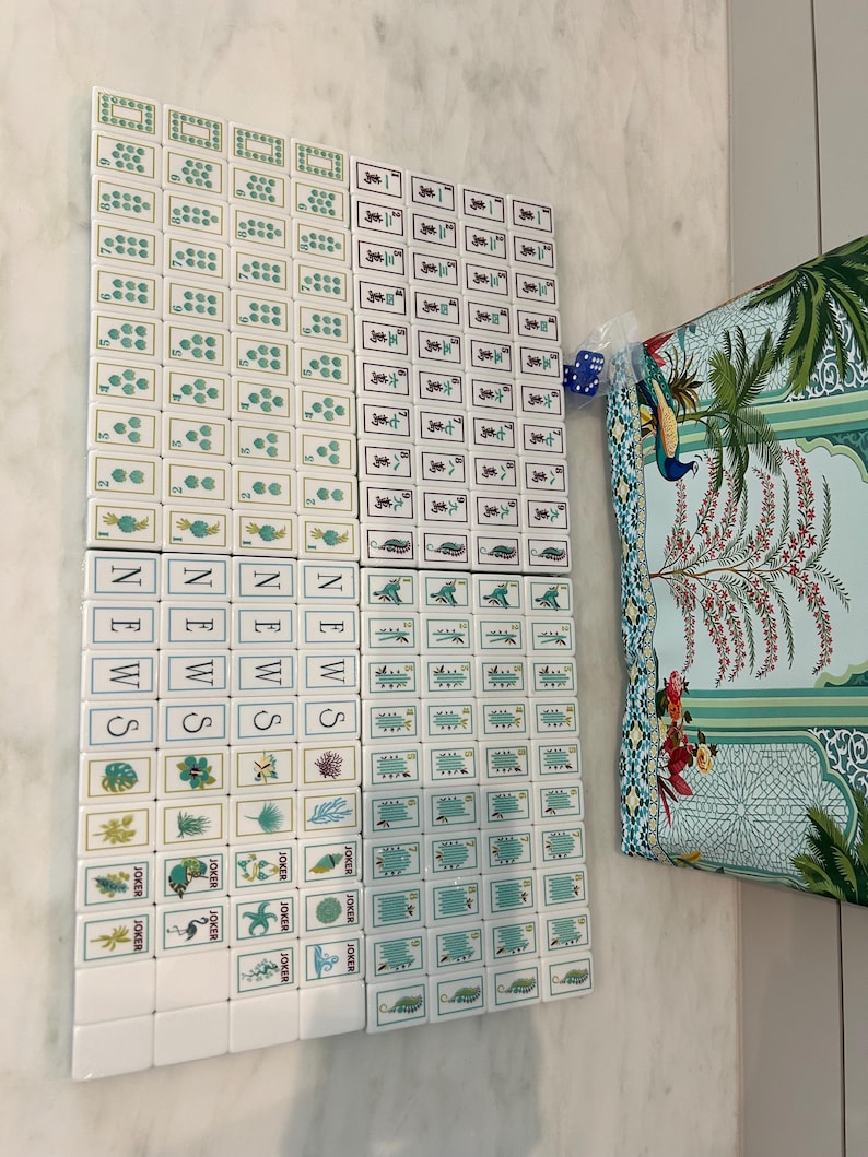 May include: A set of white mahjong tiles featuring green and black ink illustrations of numbers, symbols, and nature motifs, including tiles labelled NEWS and JOKER. A blue die and a patterned fabric bag with a peacock and tree design are included.