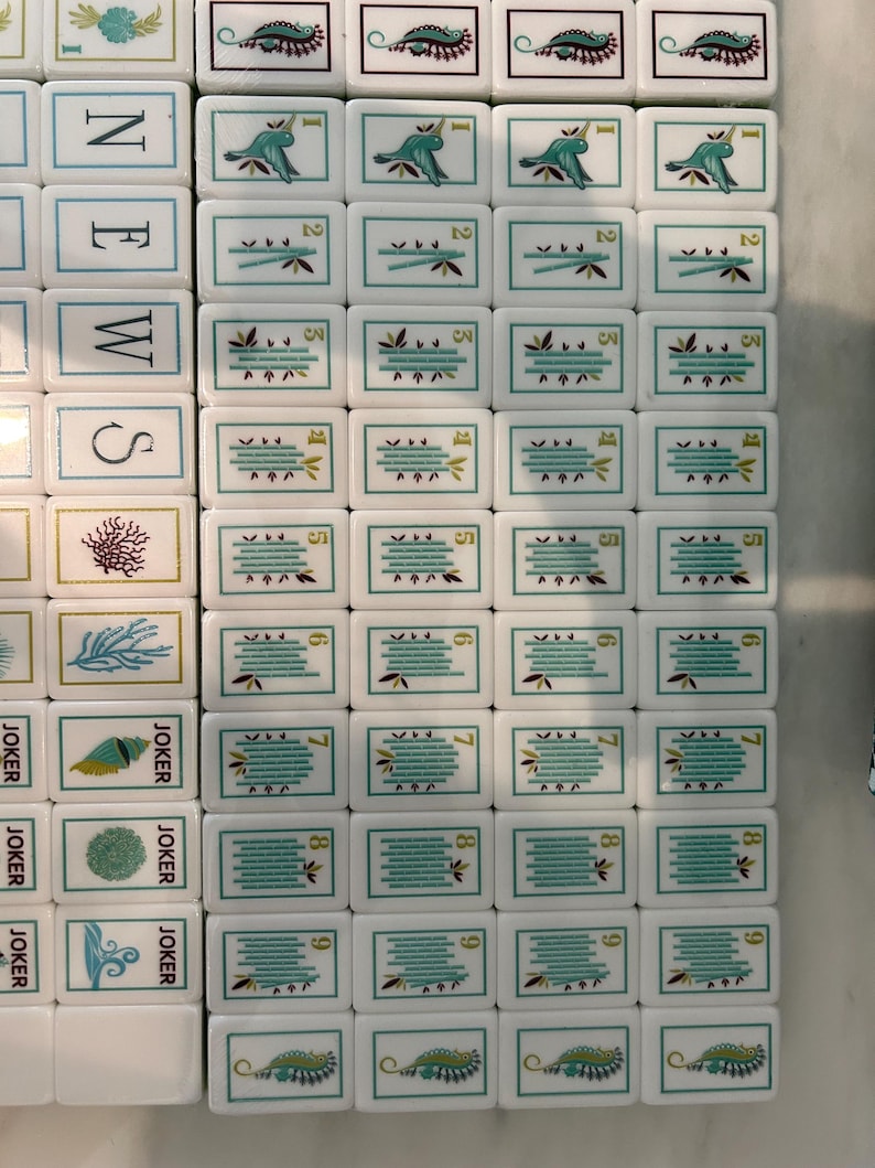 May include: A collection of white rectangular Mahjong tiles arranged in a grid. The tiles feature various illustrations including hummingbirds, bamboo stalks, sea life, and the word NEWS vertically. Each tile has a thin teal border and markings.