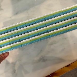May include: A rectangular stack of four rows of small, rectangular erasers wrapped in clear plastic. Each eraser features a layered design with a white top section, a lime green middle stripe, and a light blue bottom section, arranged in a grid.