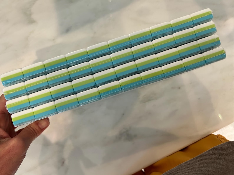 May include: A rectangular stack of four rows of small, rectangular erasers wrapped in clear plastic. Each eraser features a layered design with a white top section, a lime green middle stripe, and a light blue bottom section, arranged in a grid.