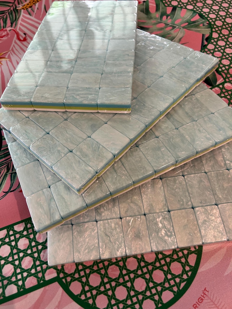 May include: A set of four rectangular mahjong tile racks stacked in a staggered formation. Each rack features a marbled light blue and white surface with a smooth, glossy finish, layered over a thin yellow and white base, all wrapped in clear plastic.