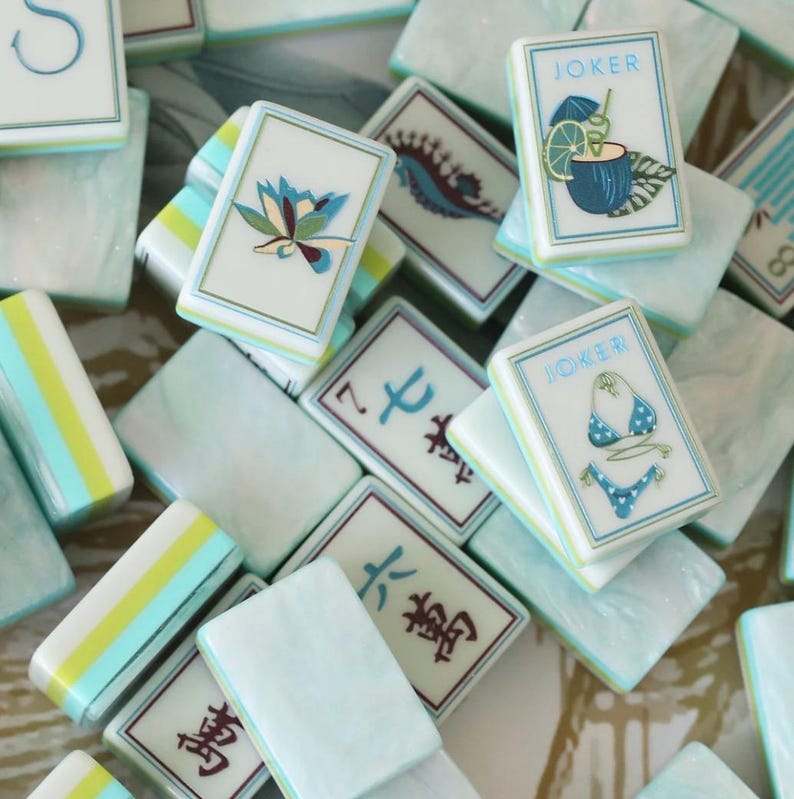 May include: A collection of rectangular mahjong tiles with a light blue marbled finish. The tiles feature various designs including a blue flower, a seahorse, a cocktail with the text JOKER, a bikini with the text JOKER, and traditional Chinese characters.