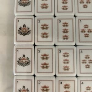 May include: A grid of white rectangular Mahjong tiles featuring a thin brown border. Each tile displays a number from one to four in the top left corner and a corresponding quantity of pink and green lotus flower illustrations centered on the face.