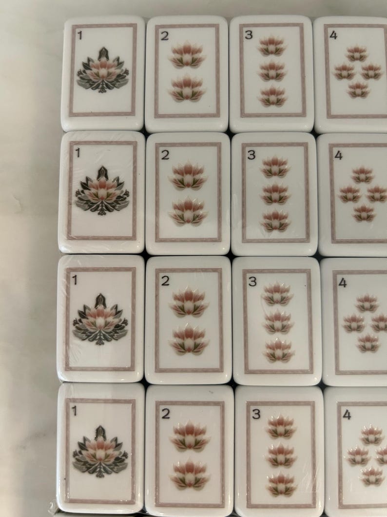 May include: A grid of white rectangular Mahjong tiles featuring a thin brown border. Each tile displays a number from one to four in the top left corner and a corresponding quantity of pink and green lotus flower illustrations centered on the face.