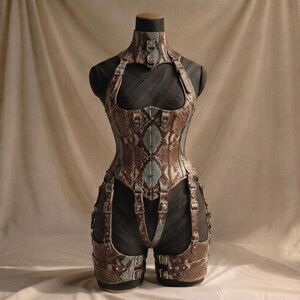 May include: A structured corset and garter set featuring a brown and teal snakeskin pattern. The garment includes a high neck collar, multiple adjustable buckled straps, and metal hardware, displayed on a mannequin over a black base layer.