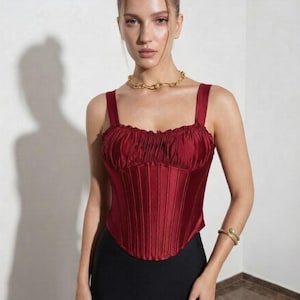 May include: A deep red satin corset top featuring a gathered, ruffled neckline, wide shoulder straps, and vertical boning details that create a structured, fitted silhouette. The garment has a curved hemline and a smooth, lustrous finish.
