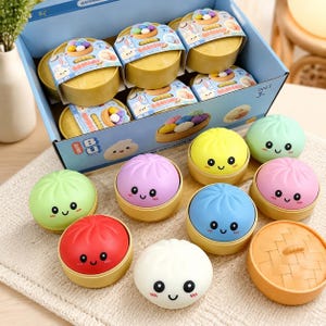 May include: A collection of round, colourful toy buns with smiling faces, each resting in a small tan plastic steamer basket. The set includes red, light green, purple, yellow, blue, and white buns, with several packaged in a blue cardboard box.