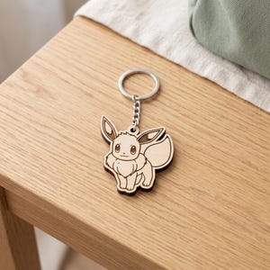 May include: A light-colored wooden keychain featuring a laser-engraved illustration of the character Eevee. The keychain includes a silver-toned metal split ring attached by a short chain, showcasing the character's pointed ears and bushy tail.