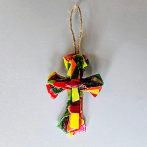 May include: A cross-shaped ornament constructed from small, irregular shards of multicoloured glass in shades of red, yellow, green, and orange. The mosaic pieces are layered to form the cross, which is suspended by a thin, light brown jute twine loop.