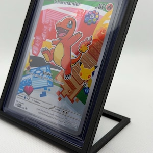 以下が含まれることがあります： 黒いプラスチックフレームに入ったヒトカゲのトレーディングカード。カードには赤とオレンジ色のヒトカゲのキャラクター、Charmanderの文字、80 HP、Emberという技が描かれています。カードの右側には小さな黄色いピカチュウのアイコンが見えます。