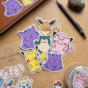 May include: A collection of vinyl stickers featuring various cartoon characters with simple, smiling faces. The stickers include yellow, purple, pink, and brown creatures, along with a small circular red and white ball sticker, all arranged on a wooden surface.