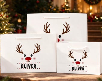 Op de afbeelding: Drie witte cadeauzakjes van verschillende formaten, elk voorzien van een bruine rendierafbeelding met een rode neus en de naam OLIVER in zwarte hoofdletters onder het rendier, afgewerkt met witte koordhengsels.