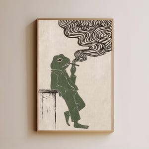 May include: A framed wall art print featuring a stylized green frog wearing a collared shirt and trousers, leaning against a table while smoking. The frog is depicted in a dark green ink style against a light beige background with swirling smoke.