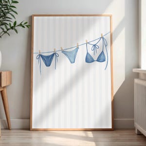 Op de afbeelding: Een ingelijste muurkunstprint met een aquarelillustratie van drie blauwe bikinionderdelen die aan een waslijn hangen tegen een achtergrond met witte en lichtgrijze verticale strepen. De set bevat twee bikinibroekjes en een bikinitop.