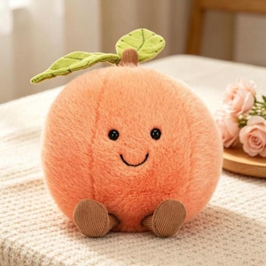 May include: A round, fuzzy, peach-colored plush toy with a smiling face, two small brown feet, and a green leaf attached to the top. The plush has a soft, textured surface and a simple, rounded shape, resembling a stylized fruit character.