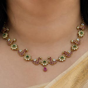 May include: A gold necklace featuring a repeating pattern of stylized peacock motifs and floral elements. The peacock shapes are inlaid with small red stones, while the floral accents contain green stones. Clear white stones border the designs.