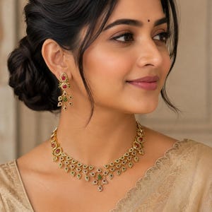Peut inclure: Un ensemble composé d'un collier et de boucles d'oreilles pendantes dorées, ornés de motifs en forme de feuilles. Les bijoux sont incrustés de petites pierres blanches transparentes, de pierres rouges vives et de pierres vertes foncées.