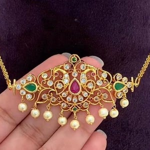 May include: A gold-toned choker necklace featuring an intricate central pendant with two peacock motifs, inlaid with clear white stones, a central teardrop-shaped pink stone, and green stone accents. The piece is finished with dangling cream-coloured pearls.