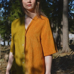 Puede incluir: Una camisa de manga corta con botones que presenta un diseño bicolor con un lado en verde oliva y el otro en amarillo mostaza. La prenda tiene un cuello en V, un dobladillo recto y un sutil patrón botánico de líneas finas estampado en la tela.