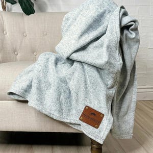 Può includere: Una coperta color grigio chiaro mélange con un aspetto morbido e testurizzato. La coperta presenta una toppa rettangolare in pelle marrone nell'angolo inferiore, impressa con il testo HERSHBERGER e una piccola icona grafica di montagna.