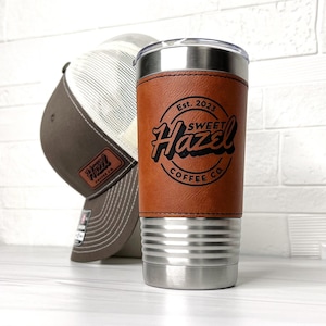 Op de afbeelding: Een reistumbler van roestvrij staal met een bruine leren omhulling. De tumbler heeft een zilveren deksel en een bruine leren omhulling met de tekst "Est. 2023 Sweet Hazel Coffee Co." De tumbler staat op een wit oppervlak naast een bruin-witte baseballpet.
