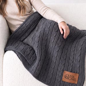 May include: A dark gray cable knit blanket featuring a rectangular tan leather patch in the bottom corner. The patch is embossed with the text OLIVIA & MICHAEL and is secured with four small metal rivets at each corner of the leather label.