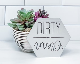 May include: A hexagonal sign with the words "Dirty" and "Clean" written on it. The sign is made of wood and has a white background with black lettering. The sign is sitting on a white countertop.
