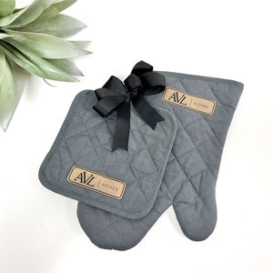 May include: A set of two gray quilted oven mitts with a brown label that says "AVL HOMES". The mitts are tied together with a black ribbon.
